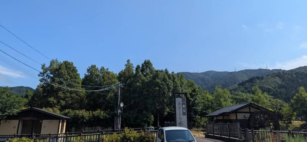 道の駅「一乗谷あさくら水の駅」の敷地から見た風景で、周囲を囲む山々と青空、そして木々に囲まれた建物が映る、自然豊かなロケーションの様子