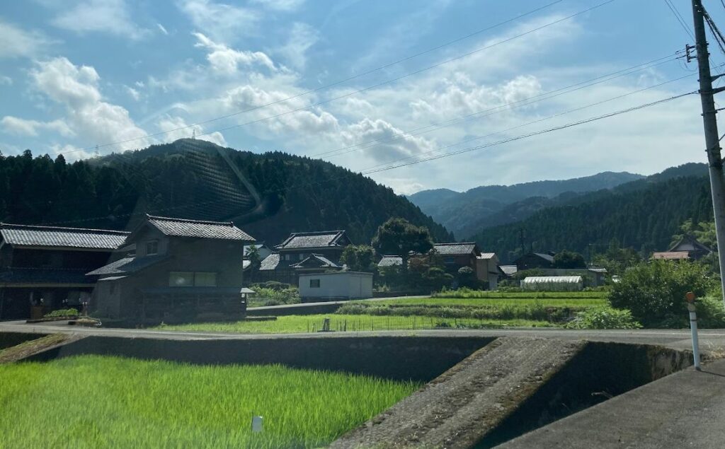 山々に囲まれた福井県の農村風景で、田んぼの奥に民家が立ち並び、のどかな集落と自然の調和が感じられる様子