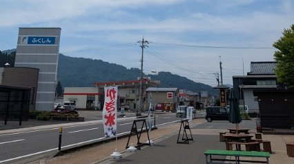 福井県池田町の「まちの駅 こってコテいけだ」周辺を撮影した写真。道沿いには「ふくしん」の看板やガソリンスタンドが見え、手前には「かき氷」ののぼり旗や木製のベンチ・テーブルが設置されている。背景には緑豊かな山々が広がり、のどかな田舎町の風景が映し出されている。