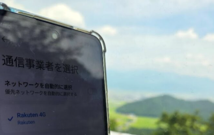 福井県大野市の国道476号線林道区間、山頂付近の開けた場所で撮影された写真。手前のスマホ画面には「Rakuten 4G」と表示されているが、実際には強力な楽天回線を掴み、珍しく山間部で「バリバリの5G」に接続できている様子が示されている。背景には山並みと田園風景が広がり、楽天回線が山奥で利用可能な非常に稀な地点であることが強調されている。