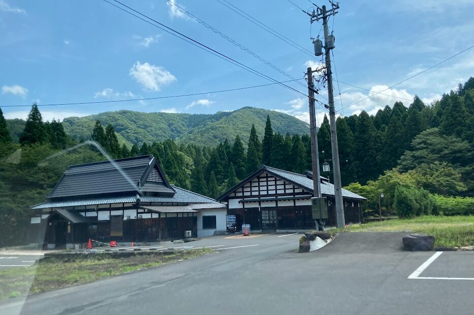 山間部にある茅葺き風の屋根を持つ古民家風建物と、木造の平屋建物が並ぶ風景写真。背後には濃い緑の杉林と山々が広がり、青空に白い雲が浮かんでいる。前景には舗装された駐車場と電柱が写り、自然に囲まれた田舎の集落の一角を切り取ったような光景となっている。
