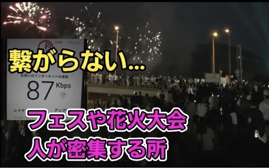 花火大会の会場で大勢の人々が密集している中、スマホの通信速度が87Kbpsまで低下し「繋がらない…」と表示されている画像。夜空に花火が打ち上がる一方で、通信が極端に遅くなる混雑環境の問題を強調している。