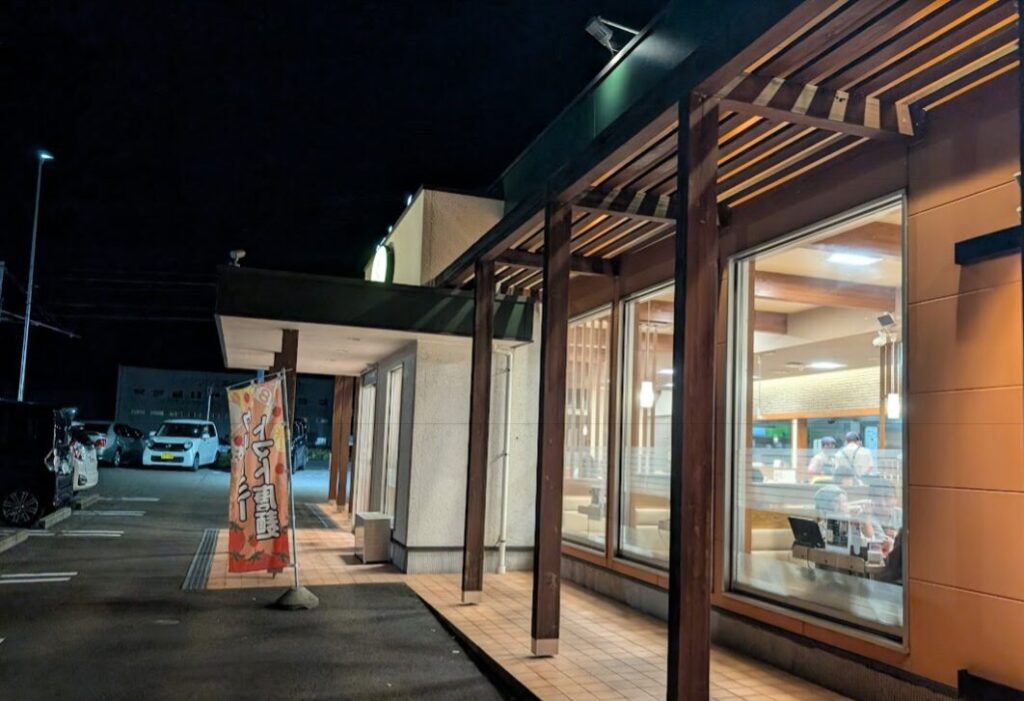 夜間に撮影されたラーメン店の外観写真。木目調の柱と大きなガラス窓が特徴で、店内では数名の客が食事をしている様子が見える。入口横には「うまいラーメン」と書かれた赤いのぼりが立ち、駐車場には複数の車が停まっている。温かな照明と夜の暗さの対比が印象的な一枚。
