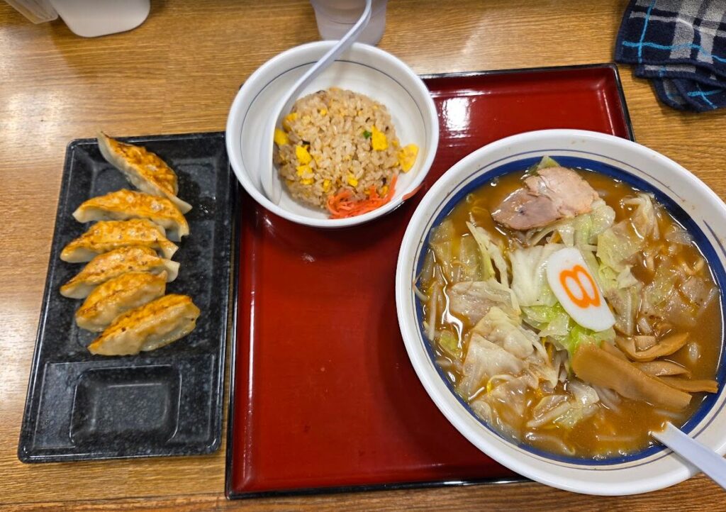 8番らーめんの定食セットを上から撮影した写真。右のどんぶりにはキャベツやチャーシュー、メンマ、なるとがのった醤油ベースのラーメン。中央の小鉢には卵とコーン、紅しょうが入りのチャーハン、左の黒皿には焼き色のついた餃子6個が並ぶ。赤いお盆の上で提供され、全体的に温かみのある照明で撮影されている。