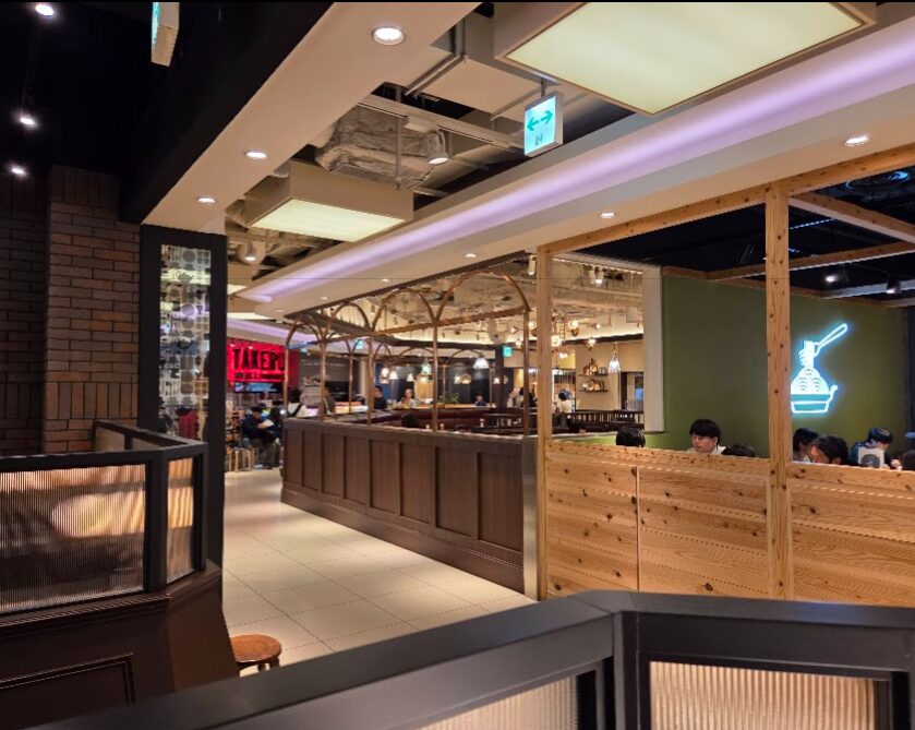 以下、**ブログ用の代替テキストのみ**を記載します。(解説なし・コピペ用)
---
店内の落ち着いた照明と木目調の仕切りが並ぶカフェ空間。奥にはカウンター席やテーブル席があり、多くの来店客が食事や会話を楽しんでいる。左奥には別店舗の赤いネオンサインが見え、天井には配管が見えるモダンなデザインが施されている。
