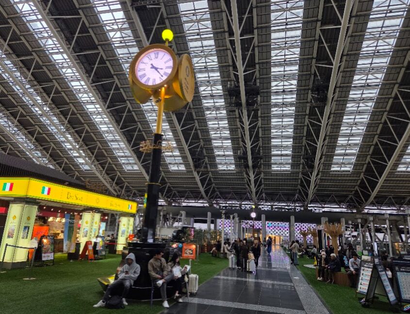 大阪駅の時空の広場の中央にある金色の大きな時計と、その周囲のベンチでくつろぐ人々。ガラス張りの高い天井から自然光が差し込み、広場には飲食店や休憩スペースが並び、行き交う人々でにぎわっている様子が写っている。