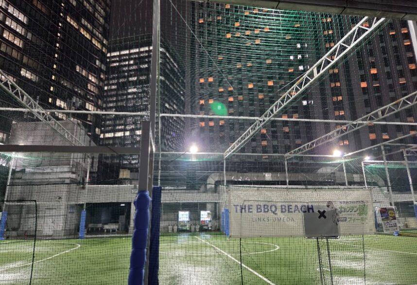 夜の梅田、ビル群に囲まれた屋上フットサルコートの風景。人工芝と照明に照らされたゴールエリア、周囲には高層ビルの窓明かりが広がり、「THE BBQ BEACH LINKS UMEDA」の幕が設置されている。