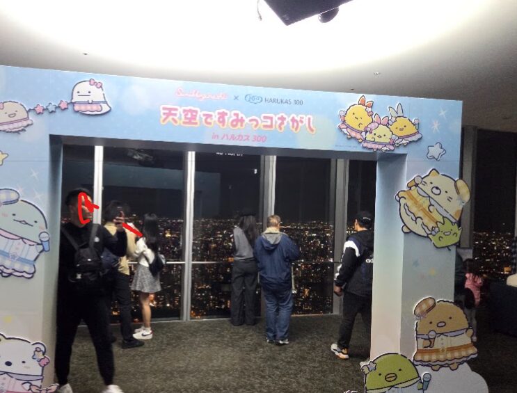 ハルカス300の展望台で開催されている「天空ですみっコさがし」のフォトスポット。カラフルなすみっコぐらしのアーチが設置され、その奥には大阪の夜景が一面に広がっている。来場者たちがガラス越しに夜景を眺めたり、写真を撮ったりしている様子が写されている。