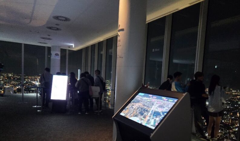 ハルカス300展望台の室内フロアで、夜景の見える窓際に多くの人が並び、大阪の街を見下ろしている様子。中央には夜景マップが映し出された案内ディスプレイが置かれ、柱や床は落ち着いたトーンでまとめられている。奥には明るい案内パネルの前に数名が集まり、夜景を楽しみながら写真を撮っている、夜景鑑賞エリアの風景。