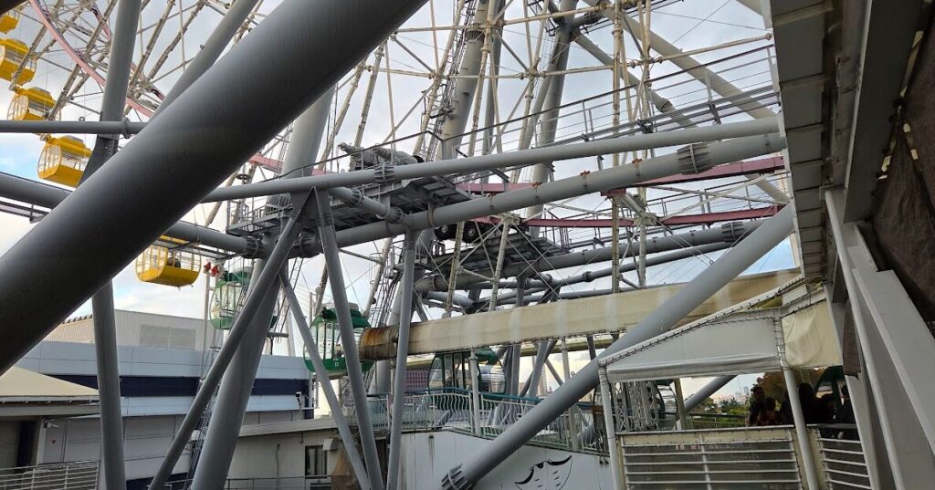 観覧車の乗り場付近を下から見上げた写真。巨大な鉄骨フレームと支柱が複雑に交差し、黄色や緑のゴンドラが周囲に配置されている。背景には青空と建物の一部が見え、観覧車の構造の迫力が強調されている。