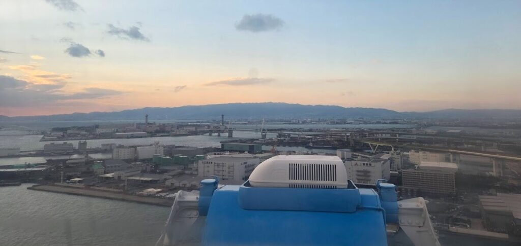 夕暮れの海遊館観覧車から見下ろす港湾エリアの風景。手前に観覧車ゴンドラ上部の青い設備が写り、その先には倉庫群や高速道路、海に囲まれた工業地帯が広がる。遠くの山並みと薄いオレンジ色の夕空が重なり、穏やかな雰囲気を作っている。