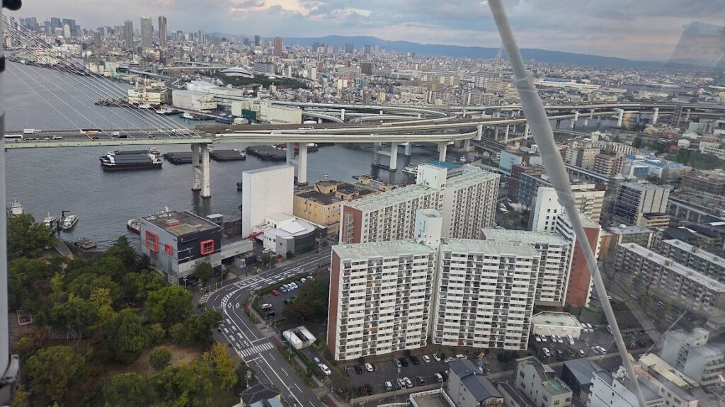 観覧車の上から大阪港エリアを見下ろした風景。手前にはマンション群が立ち並び、その奥には複雑に重なる高速道路の高架が大きくカーブしながら続いている。さらにその向こうには川と港、運搬船、倉庫群が広がり、遠景には大阪市内の高層ビル群が連なっている。夕方の柔らかい光に包まれ、都市と湾岸エリアが一望できる迫力ある眺め。
