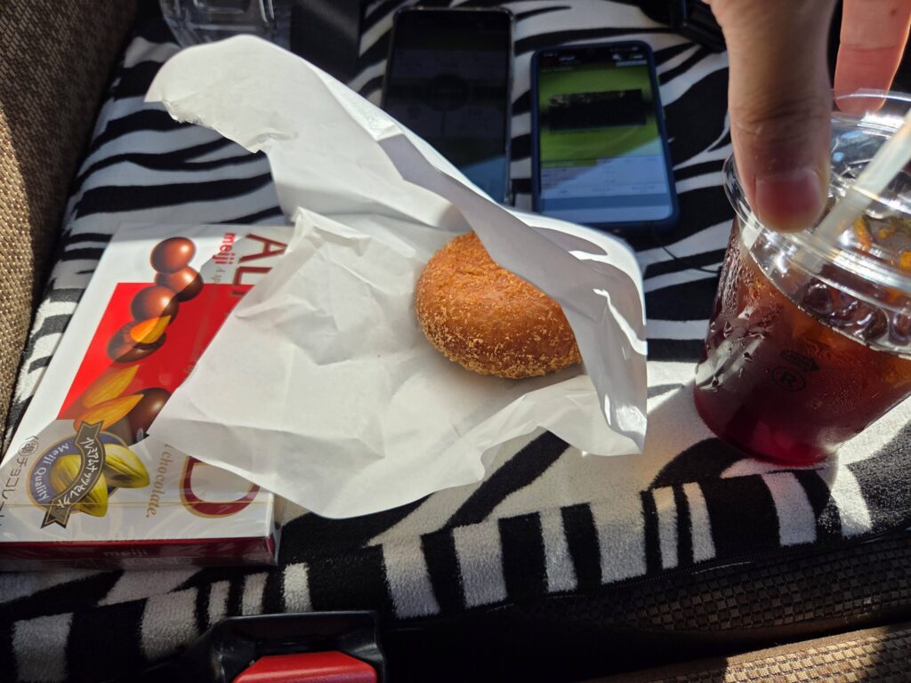 【画像7:車内での休憩風景】 ゼブラ柄のシートが特徴的な車内での休憩中の様子。手元にはカレーパンと思われる揚げパン、アイスコーヒー、明治アーモンドチョコレートが並び、奥には速度計測用と思われる複数のスマートフォンが置かれた、リアルな実測調査の合間のひとときを捉えた写真。