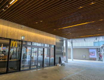 JR福井駅構内にある「マクドナルド JR福井駅店」の入り口付近の様子。木目調のルーバー天井が印象的なモダンな通路に面しており、ガラス張りの店舗外観が確認できます。この地点では平日のランチタイムに通信速度計測を実施しており、混雑する12:15時点ではahamoが230Mbps、povo 2.0が85Mbpsを記録する一方で、日本通信SIMなどのMVNO勢が1Mbpsを下回るなど、キャリアと格安SIMの速度差が顕著に表れた計測スポットです。