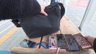 屋外の木製ベンチに5台のスマートフォンを並べ、通信速度計測の準備をしている様子。黒いショルダーバッグから端末を取り出し、実測調査を行う直前のリアルな現場風景です。この地点(JR福井駅横 ハピリン前広場)では、直後の平日12:33の計測において、povo 2.0とahamoが共に370Mbpsという高速値を記録する一方で、日本通信SIMが0.8Mbps、IIJmioが1.1Mbpsまで落ち込むなど、お昼休みの混雑による極端な速度差がデータとして観測されています。