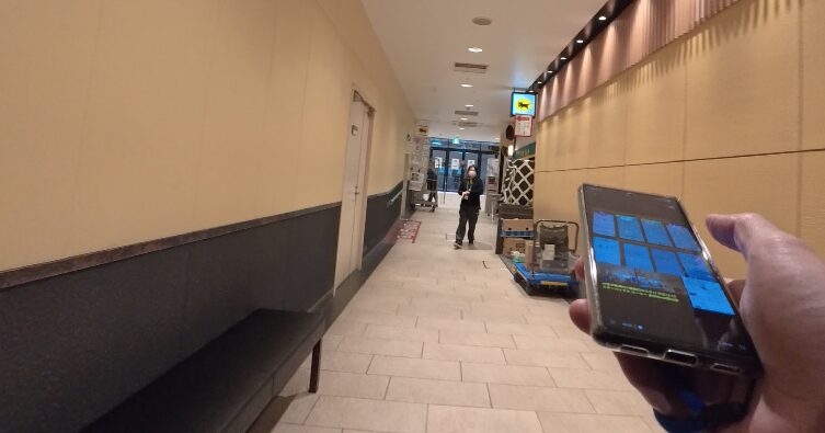 JR金沢駅構内のショッピングモール「あんと」の通路において、通信速度の測定作業を行っている様子を収めた現地写真。右手でスマートフォンを持ち、画面上には複数の通信回線の測定結果と思われるグリッド状のデータが表示されている。

背景には和風の落ち着いた壁面と、台車を押して歩く駅ビルスタッフの姿が写っており、実際に人通りのある駅施設内のリアルな環境下で測定を実施していることがわかる。前述の測定結果データが、まさにこの場所・この瞬間に取得されたものであることを証明する一次情報としての臨場感を与える画像である。