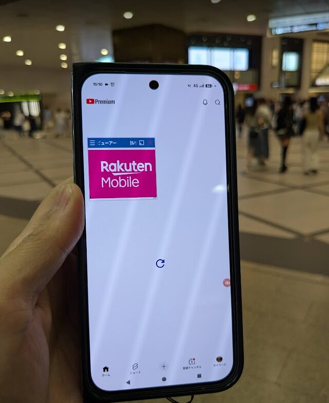 混雑した駅構内において、楽天モバイル回線でYouTubeアプリを開いているスマートフォンの画面。端末上部のステータスバーには時刻「15:10」、通信状態「4G」、バッテリー残量「86%」といった詳細な数値データが確認できるが、画面中央には青いローディングアイコンが回り続けており、通信が滞りコンテンツが読み込めていない状態が示されている。